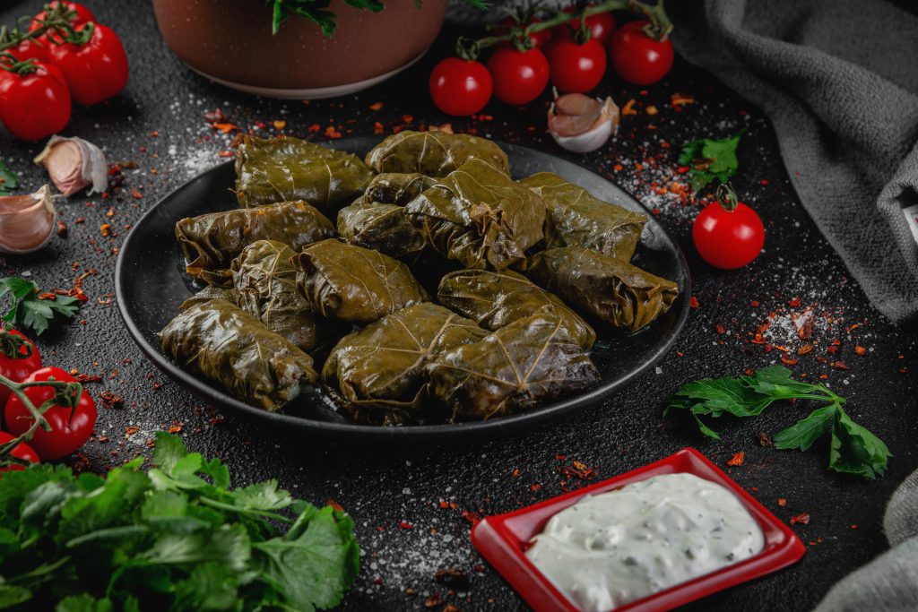 Traditional Armenian tpov dolma, stuffed grape leaves served on a dark plate with garlic matsun, tomatoes, and fresh herbs
