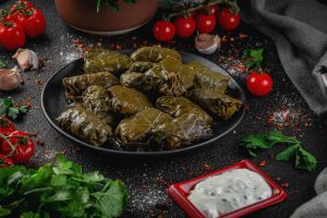 Traditional Armenian tpov dolma, stuffed grape leaves served on a dark plate with garlic matsun, tomatoes, and fresh herbs